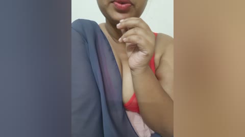 Video of a woman with medium skin tone, wearing a sheer blue robe and a red bra, her hand near her lips. Background is a gradient of brown and beige.