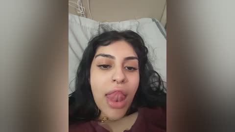 A close-up video of a young woman with long black hair, light skin, and a septum piercing, sticking out her tongue while lying on a bed with white pillows and beige walls in the background.