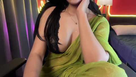 A video of a woman with long black hair, wearing a green off-shoulder dress revealing her black lace bra, sitting in a modern room with red and yellow lights and a bed in the background.