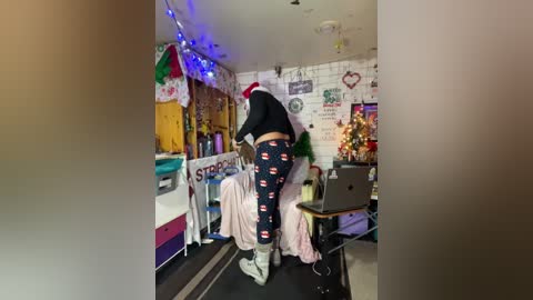 Video of a person in a festive room with holiday decorations, wearing a Santa hat, black top, and pajama pants with a Christmas theme.