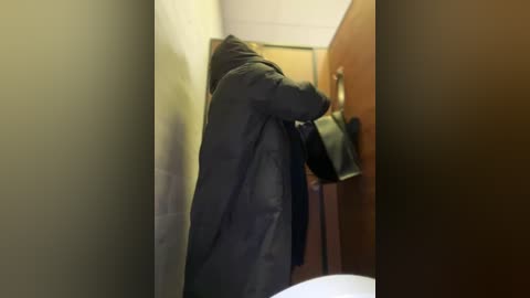 Video of a person, possibly male, wearing a dark, hooded jacket, hanging a black bag on a wall-mounted hook in a narrow, dimly lit hallway with beige walls and a wooden door.