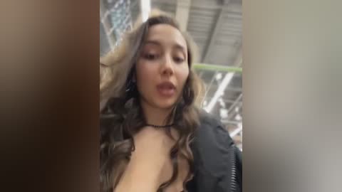 A video shows a young woman with long, wavy, brown hair and fair skin, wearing a black leather jacket and a black choker, standing in a dimly lit, industrial setting with exposed pipes and a green ladder in the background.