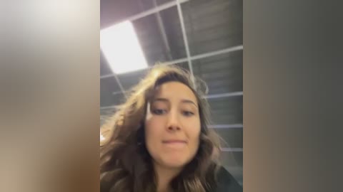 A video of a young woman with wavy brown hair, fair skin, and light makeup, smiling slightly. The background features a grid-patterned ceiling with fluorescent lights, giving a modern, industrial feel.