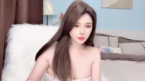 A video of a young, light-skinned woman with long, straight brown hair, wearing a white, strapless dress, sitting on a bed in a softly lit, pastel-colored bedroom.