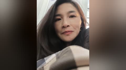 Video of an Asian woman with long, straight, dark hair, wearing a black jacket, leaning forward, looking slightly to the left. Background is blurry, likely indoor.