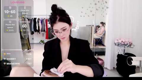 Video of a woman with glasses and dark hair tied back, wearing a black blazer, sitting at a desk, holding a pen, in a modern, minimalist room with white walls and shelves.