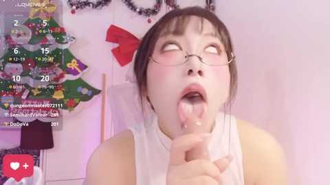 A video of an Asian woman with glasses and a white sleeveless top, licking a dildo in a festive room adorned with Christmas decorations.