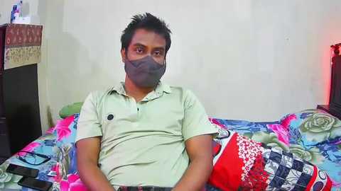 Media: Video of a South Asian man with dark skin, wearing a beige polo shirt, gray face mask, and red, patterned quilted blanket on a bed with floral-patterned bedding.