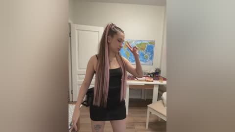 Video of a slender, light-skinned woman with long, pink-dyed braids, wearing a tight black dress, holding a phone in a bright, modern room with wooden floors, a white desk, and a world map on the wall.