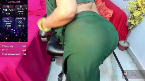 A video shows a woman in green leggings and a bra, bending over a chair, with a pink table and a potted plant in the background.