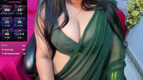 Video of a woman in a green, sheer saree with black hair, revealing cleavage, and a background of a pink chair and green plant.