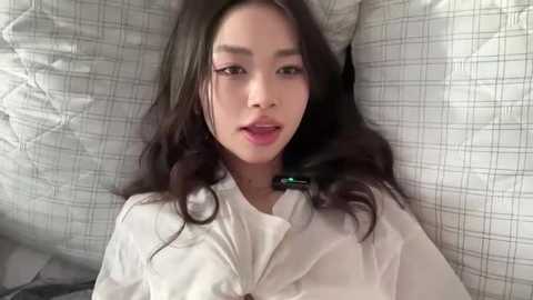 Video of an Asian woman with long, dark hair, wearing a white button-up shirt, lying on a gray plaid bedspread, with a neutral expression.