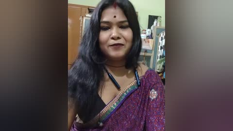 Video of an Indian woman with long black hair, wearing a purple saree, smiling, with a bindi on her forehead, indoors with shelves and a wooden door in the background.