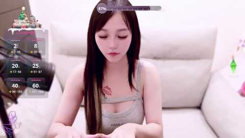 Video of an East Asian woman with long black hair, wearing a gray tank top, sitting on a white couch, with a virtual thermometer overlay and a Christmas tree in the background.