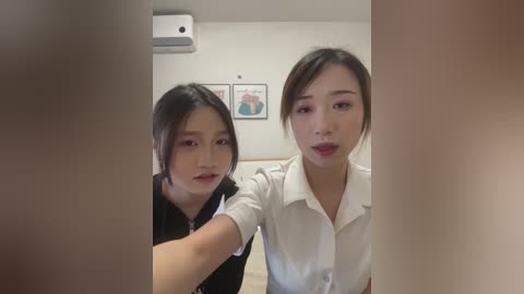 Video of two East Asian women, one with short black hair, the other with a side part, in a white blouse, standing in a minimalist room with framed artwork on a white wall.