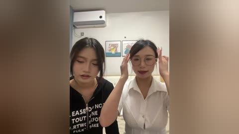 Video of two Asian women with glasses, one in a black top, the other in a white shirt, adjusting their hair in a modern office with air conditioning, framed art, and beige walls.