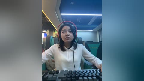Video of an Asian woman with shoulder-length black hair, wearing a white hoodie and red headphones, typing on a keyboard in a modern, dimly lit bus with teal seats.