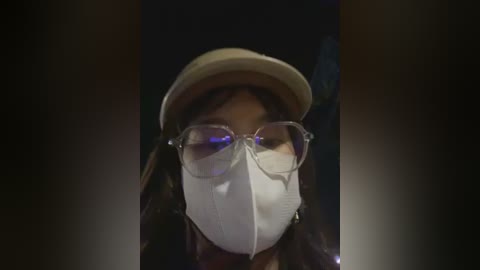A close-up video of a person with light skin wearing a beige hat, clear glasses, and a white surgical mask, set against a dark, blurry background.