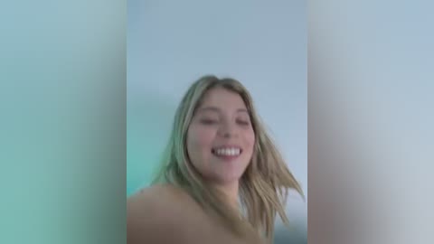 Video of a topless young woman with blonde hair and fair skin, smiling and slightly blurred, standing in front of a white background with soft lighting.