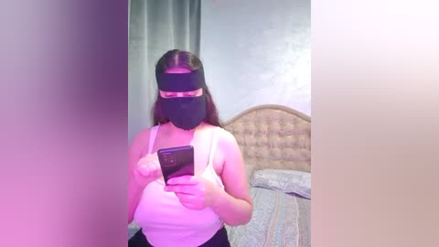 A video of a woman with long brown hair wearing a black mask and a white tank top, taking a mirror selfie in a dimly lit bedroom.