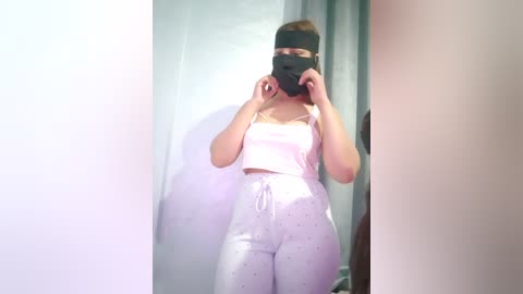 Video of a light-skinned woman with a curvy figure wearing a black face mask, light pink crop top, and high-waisted white leggings, taking a selfie in a dimly lit bathroom.