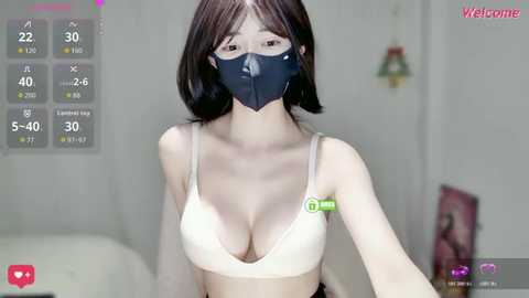 Video of an East Asian woman with fair skin and shoulder-length black hair, wearing a black face mask, white bra, and a green wristband. Background includes a white wall with a light fixture and a TV.