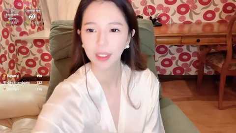 A video of an Asian woman with straight dark hair, fair skin, and light makeup, wearing a white robe, sitting on a green chair in a room with floral-patterned wallpaper and wooden furniture.