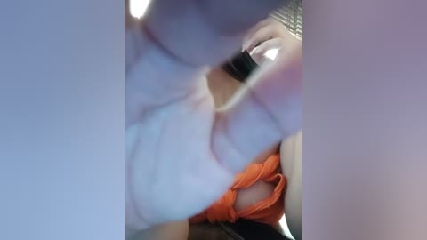 Video of a woman with fair skin, wearing a white tank top and orange shorts, holding a black cell phone, taken from a low angle, blurry background.