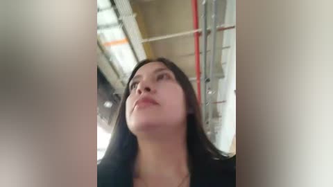 A video of a young woman with long black hair, fair skin, and a serious expression, taken from a low angle, showing her from the shoulders up. The background includes industrial pipes and a blurred figure.