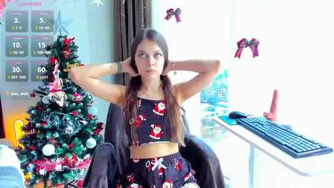 Video of a young woman in festive pajamas, hands behind head, in front of a decorated Christmas tree and keyboard, looking thoughtful.