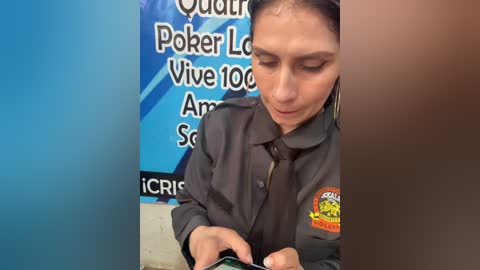 Video of a Latina woman with light brown skin and dark hair tied back, wearing a dark green uniform with a badge, focused on a computer screen in a blue and white office.