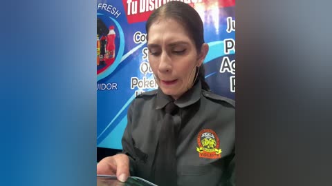 Video of a middle-aged woman with dark hair tied back, wearing a gray uniform with a badge, speaking into a microphone. Background features a blue and red promotional poster with text and images.