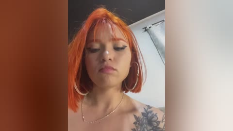 Video of a young woman with bright orange hair, wearing minimal makeup and a gold necklace, standing against a white wall with a window and curtain.