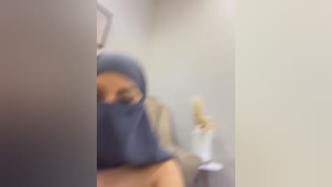 Blurry video of a woman in a blue hijab and face mask, partially visible, standing in a beige-walled hallway.