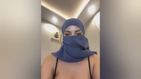 A video of a woman in a blue hijab and black spaghetti strap top, standing in a modern, dimly-lit room with white walls and recessed lighting.
