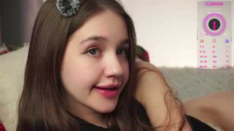 Video of a young Caucasian girl with straight brown hair, wearing a gray headband, smiling. Background shows a white wall and a pink and white digital calendar.