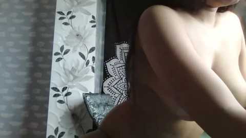 A video of a topless woman with light skin and medium-sized breasts, seen from the side, standing in a room with a floral-patterned wallpaper and a black and white mandala cushion.