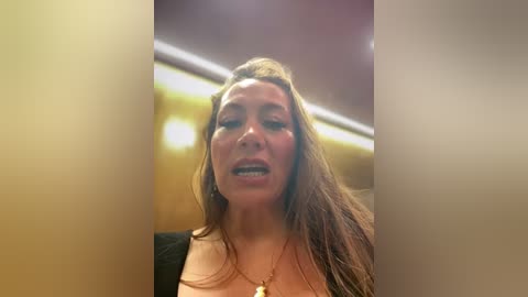 Video of a middle-aged woman with long, wavy brown hair, wearing a black top, standing in a dimly lit, beige-tiled room with a blurred background.