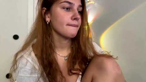 Video of a young Caucasian woman with long, wavy auburn hair, wearing a white top and a silver necklace, sitting indoors, smiling slightly, with a blurred background.
