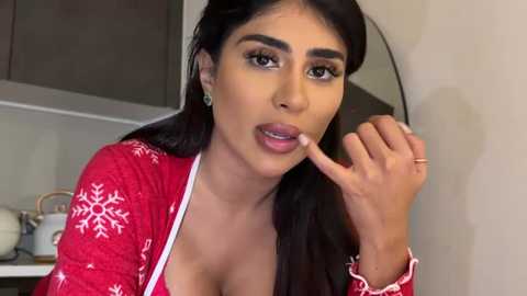 Video of a young woman with long dark hair, tan skin, and large breasts, wearing a red sweater with white snowflakes, eating a finger, in a modern kitchen with white cabinets and a round mirror.