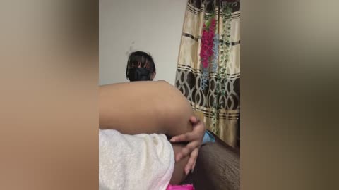 Video of a woman with medium skin tone, wearing a black mask, lying on a bed, legs spread, with a floral-patterned curtain in the background.