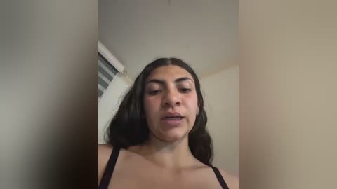 Video of a young woman with long, dark hair and light skin, wearing a black bra, standing in a beige room with a window partially visible.