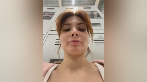 A video of a young woman with light skin and auburn hair, slightly smiling, wearing a pink hoodie, in a locker room with white lockers and posters on the wall.