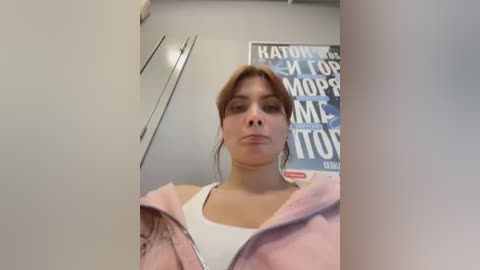 Video of a young woman with fair skin, brown hair, and light makeup, wearing a white tank top and pink jacket, standing in a modern, white-walled room with a Russian text poster.