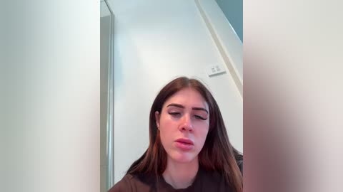 Video of a young woman with long brown hair, wearing a black shirt, sitting in a modern, minimalistic room with white walls and a glass door.