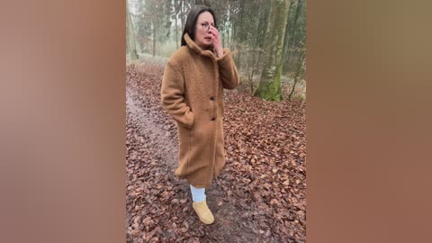 Video of a woman with shoulder-length hair, wearing a brown coat, blue socks, and tan boots, standing on a gravel path surrounded by autumn leaves.