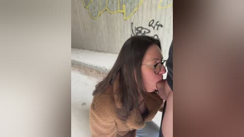 A video shows a young woman with long brown hair, glasses, and a brown sweater performing oral sex on a man in a graffiti-covered, dimly-lit bathroom.