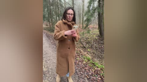 Video of a woman with glasses and brown hair, wearing a long brown coat and beige boots, holding a white coffee cup, standing on a dirt path in a forest with bare trees and green shrubs.