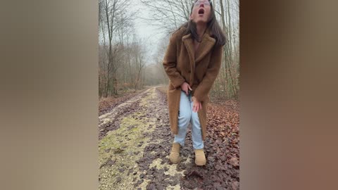 Video of a woman in a brown coat, light blue jeans, and tan boots, bending over with her hands on her knees, laughing on a muddy, leaf-strewn forest path.