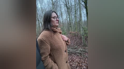 Video of a middle-aged woman with glasses and long brown hair, wearing a brown coat, standing in a leaf-covered forest, looking thoughtful.